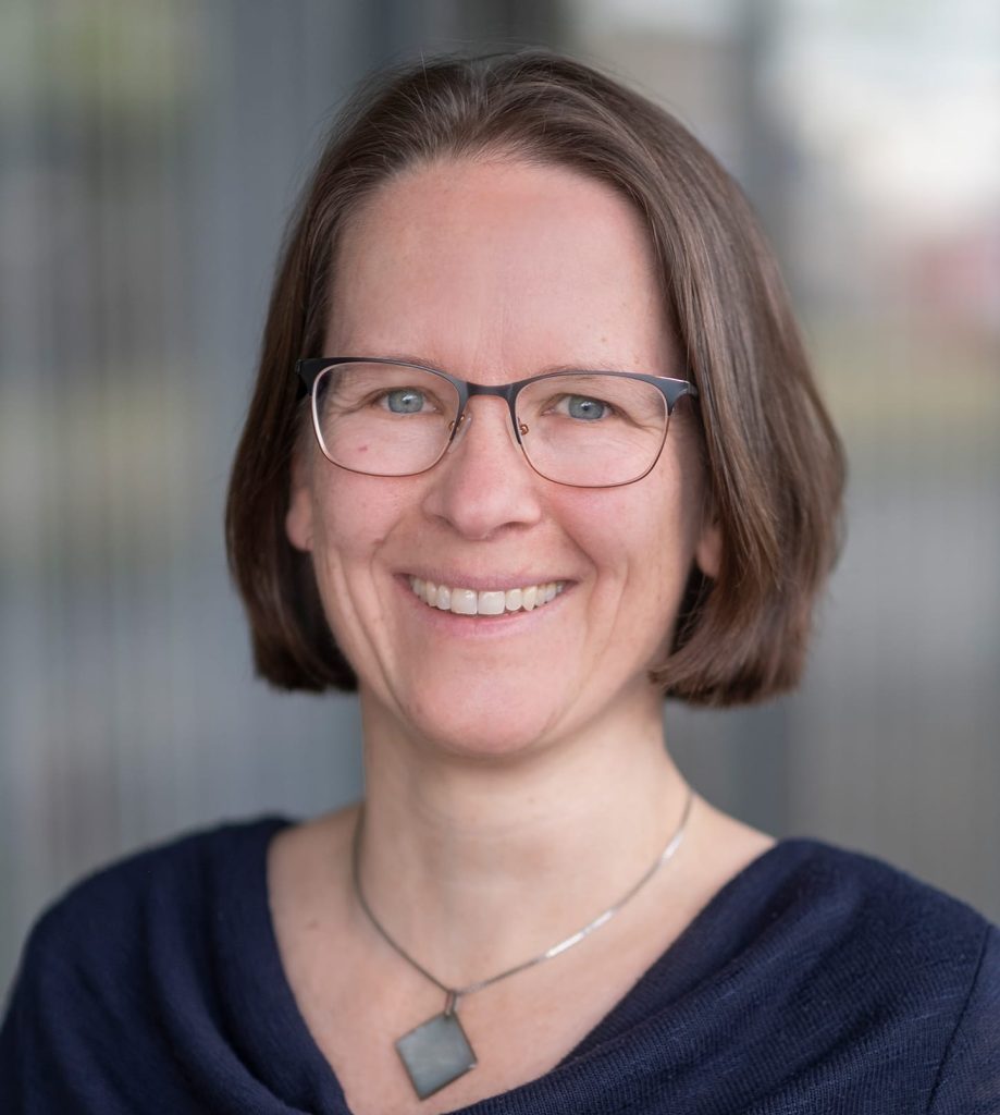 Das Foto zeigt eine Frau mit heller Haut und kinnlangen braunen Haaren. Sie trägt eine Brille und eine Kette. Sie hat einen blauen Pullover an und lacht in die Kamera.