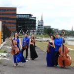 Das Bild zeigt einige Musikerinnen und Musiker mit ihren Streichinstrumenten auf einem Weg. Sie tragen elegante schwarze und blaue Kleidung. Im Hintergrund sind Gebäude zu sehen.