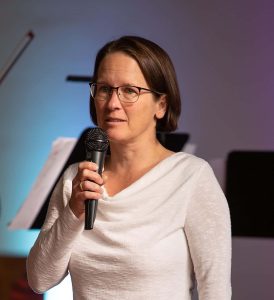 Konzertmoderation ( Einfache Sprache) Katrin Anders in weißem Oberteil und schwarzer Hose spricht in ein Mikrofon.