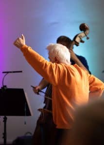 Im Hintergrund musiziert ein Kontrabassist. Er wird fast vollständig von einem Mann mit weißen Haaren und orangefarbenem Pullover verdeckt. Der Mann läuft durch das Bild und streckt seinen linken Arm nach oben.