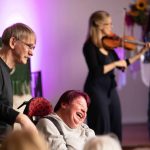 Im Hintergrund musiziert eine Geigerin. Vorn im Bild schiebt ein Mann eine lachende Frau im Rollstuhl.