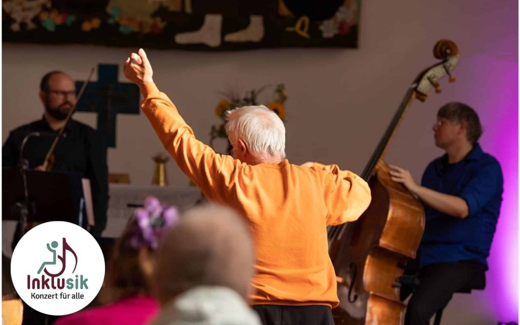 Im Hintergrund stehen zwei Musiker vor einem Altar, rechts im Bild ein Kontrabassist. Vor ihnen steht ein Mann mit weißen Haaren und orangefarbenem Pullover und schaut sie an. Er streckt seinen linken Arm nach oben.