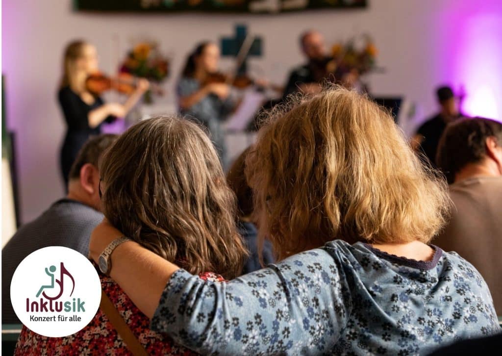 Zwei Frauen, nebeneinander sitzend und von hinten zu sehen, in einem Konzert mit Streich-Instrumentalist*innen. Die rechte Frau hat ihren linken Arm um die Schulter der linken Frau gelegt.